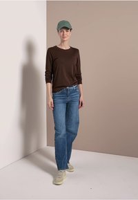 Bruine lange mouwen top, blauwe losvallende jeans en lichte sneakers. Accessoire is een groene pet. Neutrale achtergrond met zachte schaduwen.