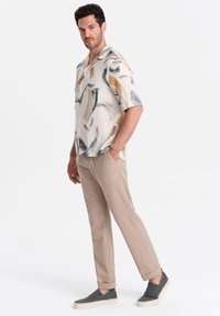 Camicia a maniche corte con motivi astratti beige e blu, abbinata a pantaloni khaki chiari e scarpe slip-on grigie con suole in gomma.