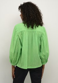 Green, dotted blouse with puff sleeves, textured fabric, and a gathered back design. Styled with dark trousers. Back view.