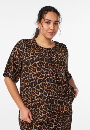 Ung kvinde iført kortærmet skjorte med leopardmønster og knapper, med den ene hånd i lommen, stående mod en hvid baggrund.