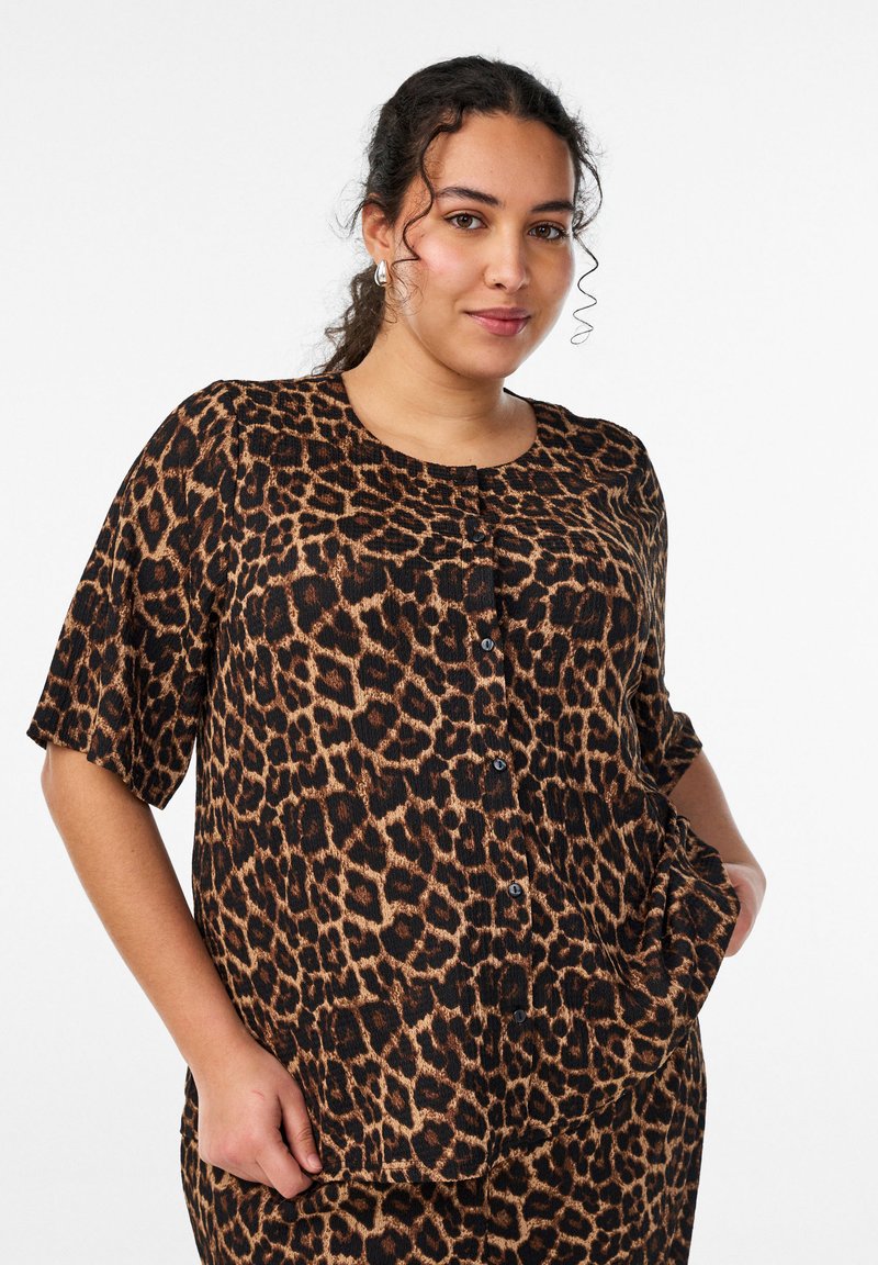 Jauna sieviete, tērpusies īsām piedurknēm apdrukātā pogas krekliņā ar leoparda rakstu, viena roka kabatā, stāvot pret baltu fonu.