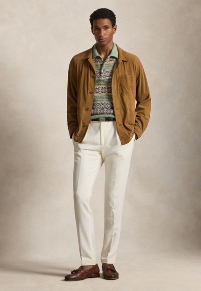 Uomo con giacca marrone, maglione verde lavorato a maglia con motivo, pantaloni bianchi e mocassini in pelle marrone con nappine, in piedi davanti a uno sfondo neutro.