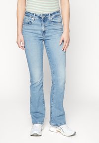 Personne portant un jean bootcut bleu clair, un haut rayé et des baskets blanches, se tenant contre un fond clair.