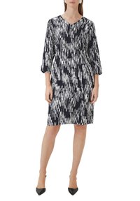 Frau in einem knielangen Kleid mit abstraktem schwarz-weißen Muster und Dreiviertelärmeln, dazu schwarze spitze High Heels.