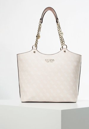 Beige värinen Guess-käsilaukku, jossa on kullanväriset ketjukahvat, logokuvio ja etuosan keskellä kohokuvioitu teksti "GUESS EST 1981".