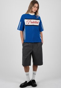 Mujer que lleva una camiseta azul de manga corta con cremallera y texto "Prohibido", pantalones cortos grises de rayas finas, calcetines blancos y mocasines negros, de pie con las manos en los bolsillos.