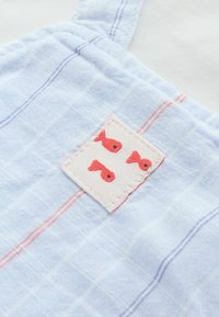 Lyseblåt stof med tynde lilla og lyserøde striber, med et lille mærke syet på med tre røde fiskeikoner.