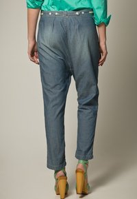 Jeans in denim di un blu medio con un design affusolato, caratterizzati da pieghe in vita e chiusura con bottone. Abbinati a sandali con tacco verde.