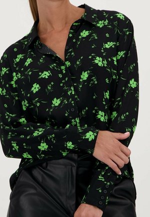 Kobieta ubrana w czarną koszulę na guziki z jasnym zielonym kwiatowym wzorem, w połączeniu z czarnymi skórzanymi spodniami, z założonymi na sobie ramionami.