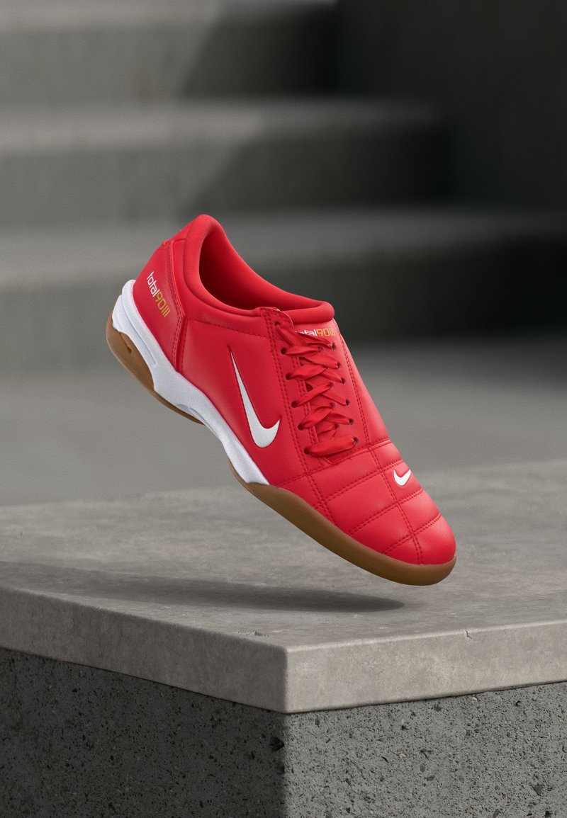 Κόκκινο εσωτερικό παπούτσι ποδοσφαίρου Nike με λευκό λογότυπο swoosh, γλωσσικό πάτο από καουτσούκ και σήμανση "total90 III", αιωρούμενο πάνω από μια τσιμεντένια άκρη.
