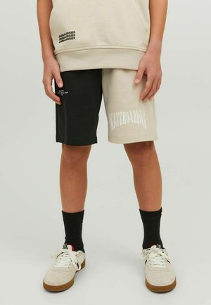 Pantaloni corti neri e beige con la scritta "SKATEBOARDING" sul lato beige. Abbinati a un hoodie crema e sneakers bianche. Tessuto in cotone misto testurizzato.