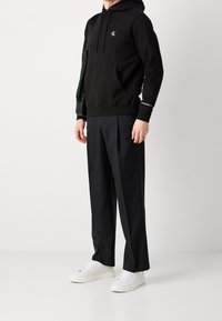 Black cotton hoodie with front pocket and logo, paired with black pleated trousers and white sneakers. Adjustable straps on the hoodie.