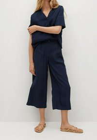 Navy dvoudílný outfit složený z topu s krátkým rukávem a výstřihem do V a širokých kalhot, vyrobený z hladkého materiálu, doplněný světlými plochými sandály.