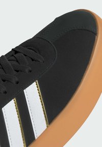 Crne tenisice s glatkom gornjom površinom, s bijelim prugama i zlatnim akcentom. Zaobljen vrh, teksturirane vezice i guma boje gume.