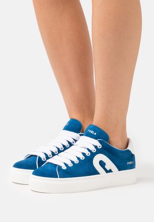 Eine Person trägt blue und weiße Furla Sneaker mit weißen Schnürsenkeln und steht vor einem einfarbigen weißen Hintergrund.