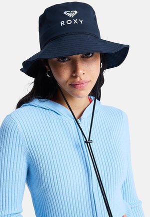 Young woman wearing a black Roxy bucket hat with strap and a light blue ribbed zip-up hoodie against a white background.