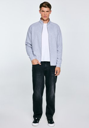 Młody mężczyzna stojący z jedną ręką w kieszeni, ubrany w jasnoszarą kurtkę na zamek, białą koszulkę, czarne dżinsy oraz czarne trampki z białymi podeszwami.