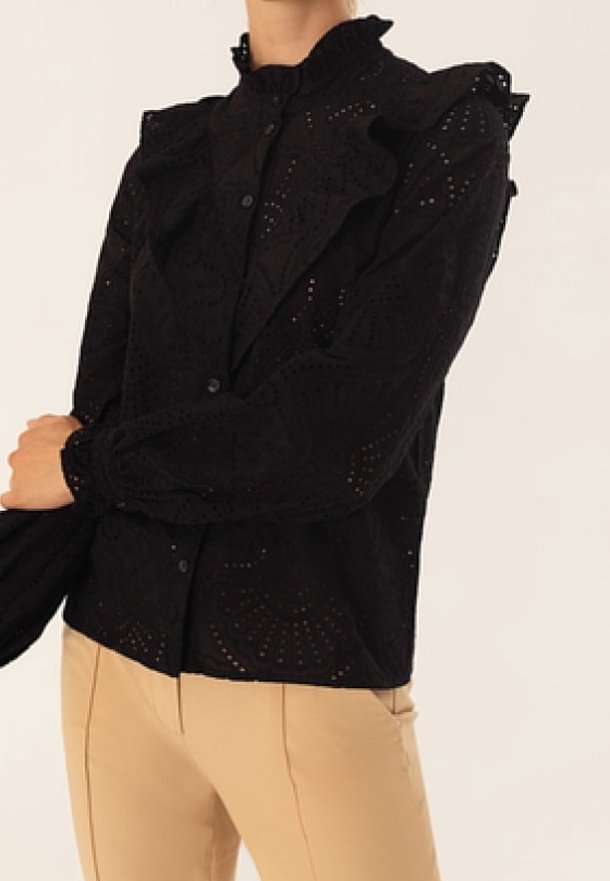 Blouse noire à manches longues avec détails œillet, col et épaules à volants, associée à un pantalon beige sur une personne croisant les bras.