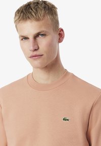 Sudadera de algodón color melocotón claro con cuello redondo, que presenta un pequeño logotipo de cocodrilo verde en el lado izquierdo del pecho. Textura suave y diseño sencillo.