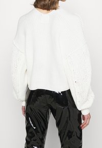 Grob gestrickter weißer Pullover mit Ballonärmeln, gerippter Textur und locker gewebten Details. Kombiniert mit glänzenden schwarzen Lackhosen.