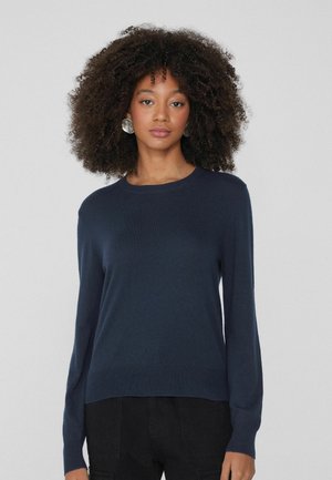 Maglione blu navy in maglia morbida, con collo a giro, maniche lunghe e polsini e orlo a coste, indossato con pantaloni neri.