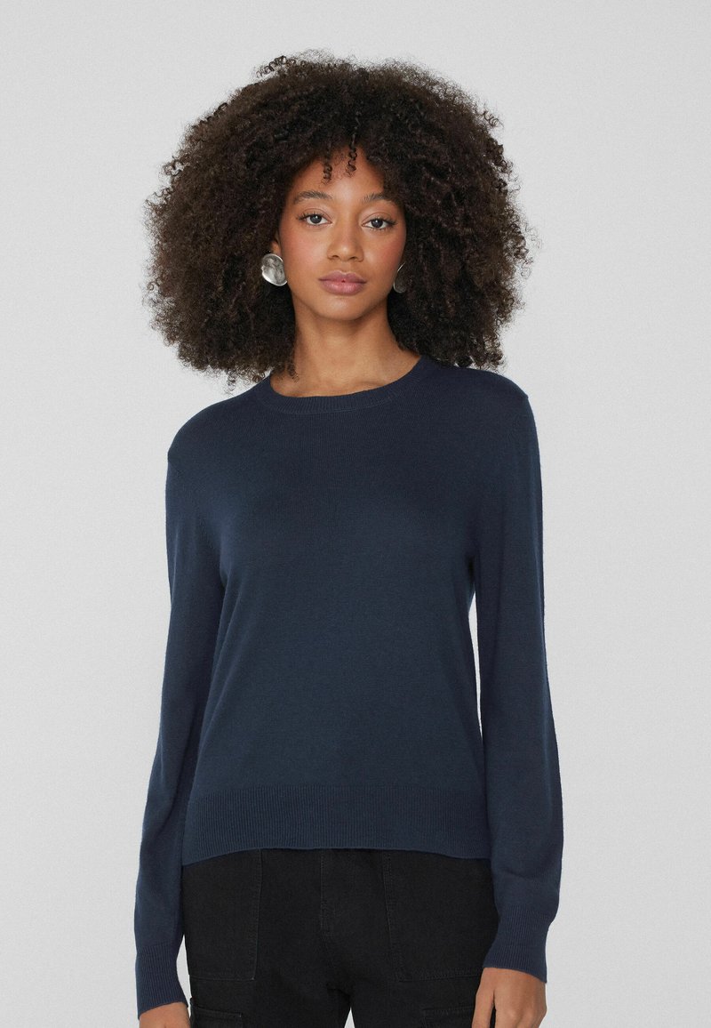 Dunkelblauer Pullover aus weichem Strick, mit Rundhalsausschnitt, langen Ärmeln sowie gerippten Bündchen und Saum, getragen zu schwarzen Hosen.