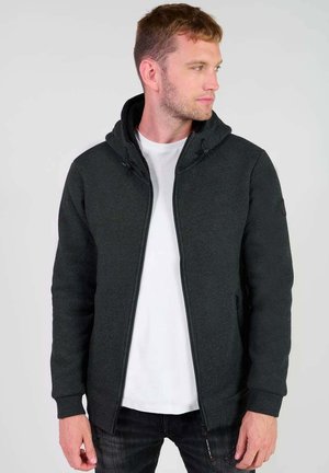 Sweatshirt à capuche noir en tissu texturé, avec une fermeture éclair à l'avant, une capuche avec cordon de serrage et des poches latérales, porté par-dessus un T-shirt blanc.