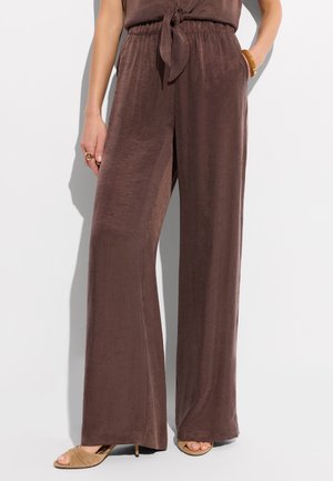 Kobieta nosząca szerokie brązowe spodnie z elastycznym pasem i wiązanym detalem, połączone z beżowymi sandałami na obcasie, na białym tle.