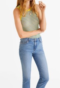 Mujer vestida con jeans de cintura alta azul y una blusa sin mangas de punto amarillo, verde y azul con un patrón, de pie con un brazo cruzado.