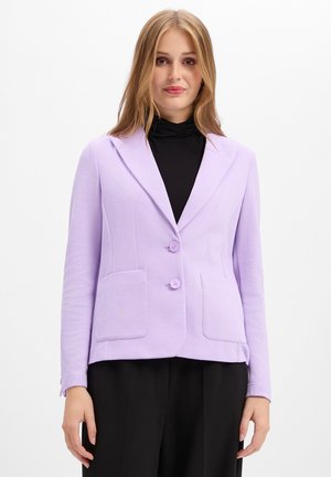 Junge Frau steht, trägt ein hellviolettes Sakko mit zwei Knöpfen über einem schwarzen Top und schwarzen Hosen vor einem weißen, einfarbigen Hintergrund.
