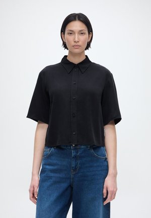Femme aux cheveux courts et foncés portant une chemise ample noire à manches courtes boutonnée et un jean bleu, sur un fond clair uni.