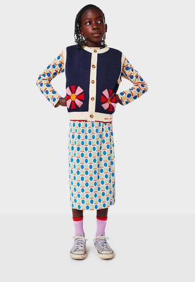 Knit cardigan in navy with cream trim, floral appliqués, and buttons. Patterned, knee-length skirt featuring blue and orange circles. Silver sneakers.