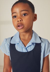 En ljusblå skjorta med krage och puffärmar under en mörk marinblå hängselklänning, med plisserade detaljer på skjortan.