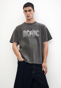 Siva bombažna majica s kratkimi rokavi in sproščenim krojem, ki ima bel logo AC/DC. V kombinaciji z temnimi, ohlapnimi kavbojkami.