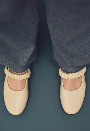 Beige nahka kengät pyöreillä varpailla ja joustavalla frilla-yksityiskohdalla nilkassa, yhdistettyinä harmaisiin housuihin ja tekstuuriseen taustaan.