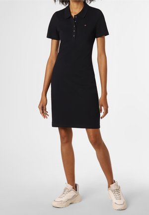 Kvinde iført en tætsiddende sort kortærmet polokjole kombineret med chunky off-white sneakers, stående mod en ensfarvet baggrund.