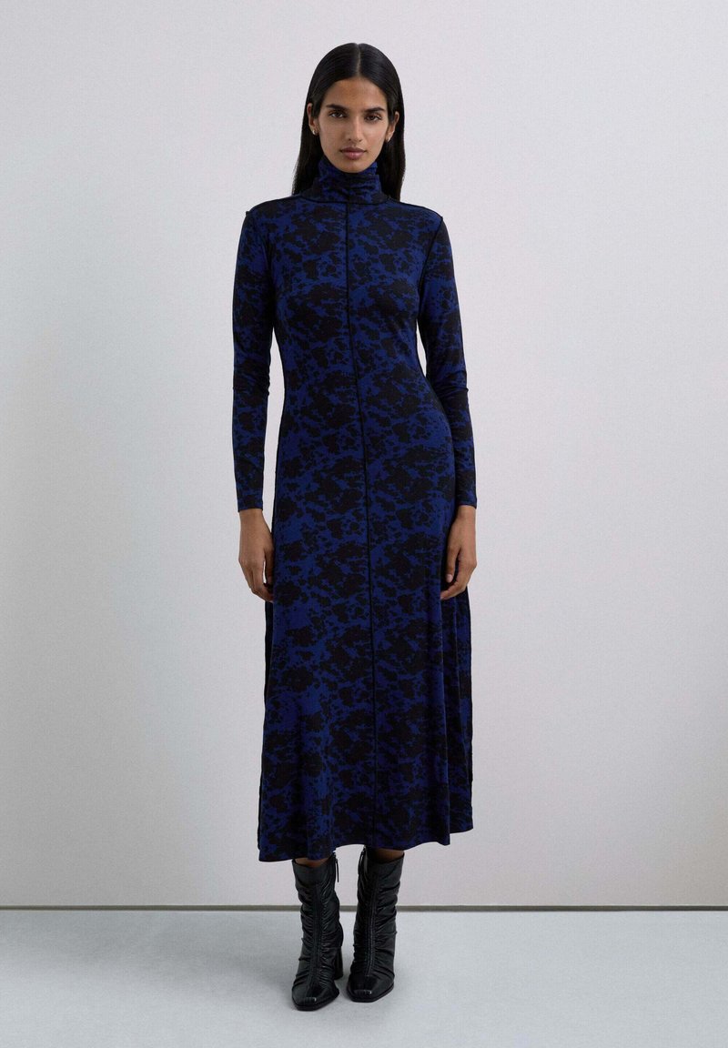 Abito a maniche lunghe e collo alto di colore blu scuro con motivo astratto nero, silhouette aderente, realizzato in tessuto elasticizzato, abbinato a stivali neri.