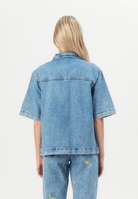 Jachetă din denim de culoare albastru deschis, croială relaxată, cu mâneci scurte, guler clasic și detalii de cusătură minime. Purtați-o împreună cu pantaloni din denim brodați.