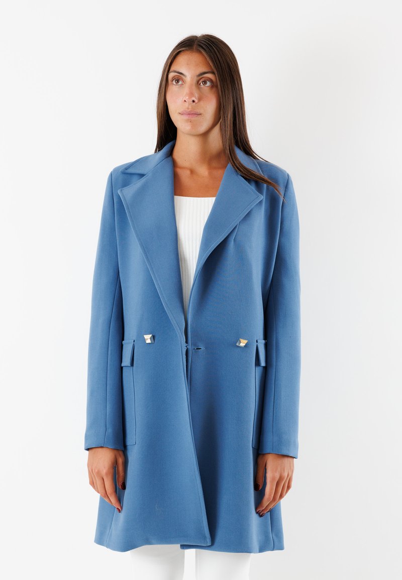 Cappotto lungo blu con colletto strutturato, tasche laterali e dettagli metallici. Tessuto liscio e vestibilità morbida, che arriva a metà coscia.