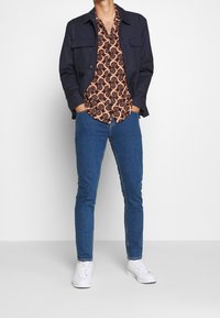 Chaqueta azul marino sobre una camisa marrón y negra con estampado de remolinos, combinada con vaqueros azules y zapatillas blancas.