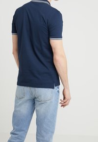 Camisa polo de color azul marino con dos franjas blancas en el cuello, manga corta y aberturas laterales. Combinada con jeans de mezclilla azul claro. Modelo visto desde atrás.