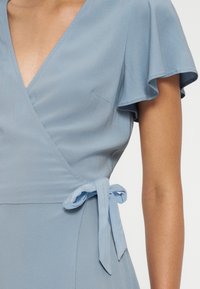 Robe portefeuille bleu clair avec manches courtes volantées et un nœud noué à la taille portée par une personne à la peau de ton moyen