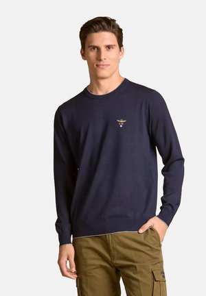 Uomo che indossa un maglione a maniche lunghe blu navy con un piccolo emblema sul petto e pantaloni cargo verdi oliva, in piedi con una mano in tasca.