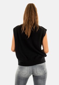 Suéter negro sin mangas con dobladillo acanalado, hecho de material suave. El modelo lleva puesto unos jeans ajustados grises, mostrando un look casual desde atrás.