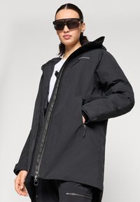 Chaqueta impermeable negra con capucha, frente con cremallera y bolsillos laterales. Presenta una textura suave y un diseño minimalista.