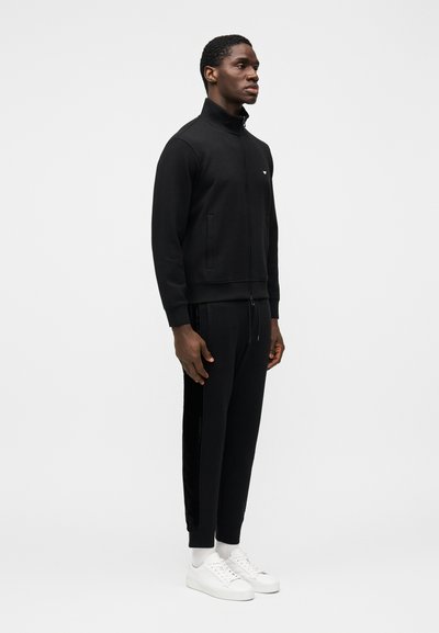 Juoda džemperis su užtrauktuku, aukštu apykakliu ir priekinių kišenių, derinama su atitinkančiomis bėgiojimo kelnėmis su šoninėmis juostelėmis. Aprangą papildo balti sportbačiai.