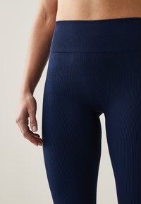 Marineblå ribbed leggings med høj talje, lavet af strækbart materiale, med midt på læggen længde og glat tekstur. Hånd hvilende på benet.