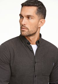 Camicia button-up marrone scuro con colletto, dotata di taschino sul lato sinistro del petto, tessuto testurizzato e cuciture bianche a contrasto sullo sprone.