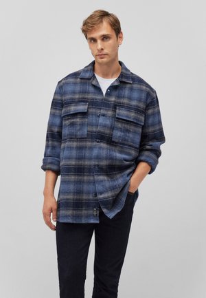 Blue plaid flannel shirt featuring two chest pockets, button closure, and long sleeves, paired with dark pants. Smooth texture and relaxed fit.