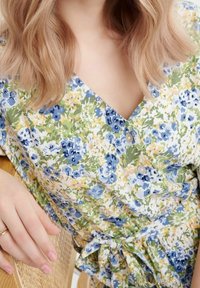 Květinové šaty s obalovým stylem s modrým, zeleným a žlutým vzorem. Výstřih do V s knoflíky, páskový detail na uvázání a lehká látka.